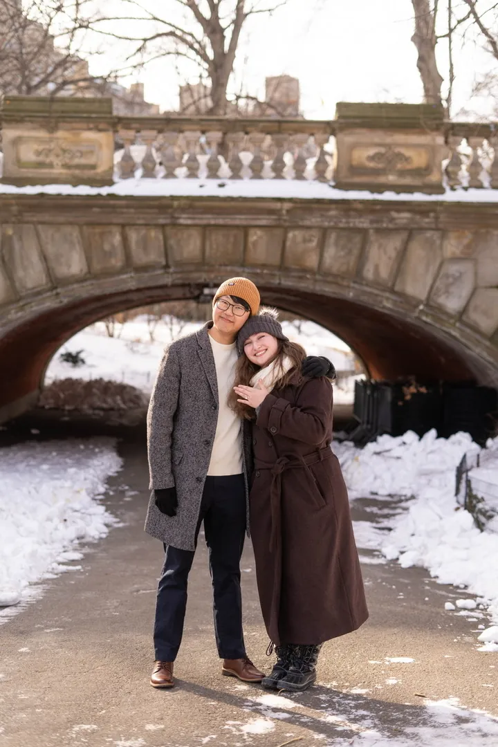 Hannah and Daniel in winter coats on a snowy path under a stone bridge in the park, her hand on his chest with the new ring on it